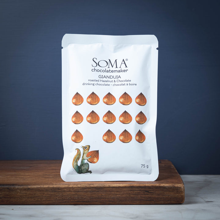 Package of SOMA Gianduja roasted hazelnut drinking chocolate on a wooden board with a dark blue background