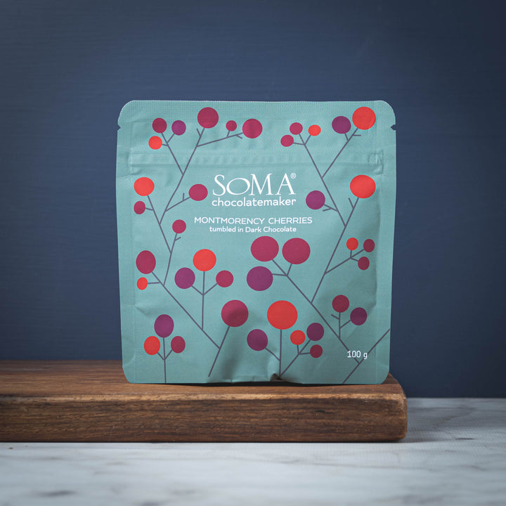 SOMA Montmorency Cherries Tumbled in Dark Chocolate packaging with cherry design on a wooden board against a dark blue background
