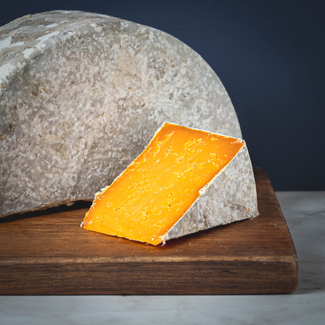 A wedge of orange Red Leicester cheese on a wooden board with a half wheel in the background
